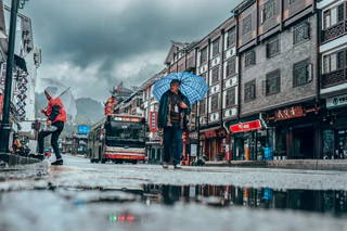 Two men carrying umbrellas cross a rain-wet street as a bus passes behind them — UNSTAGED · SERIE /001 · MOMENT /006 · ARCHIVE ID S001-M006 · SOURCE L013-M006.