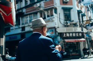 An older man in a checked hat exhales cigarette smoke on a city street — UNSTAGED · SERIE /001 · MOMENT /003 · ARCHIVE ID S001-M003 · SOURCE L013-M002.