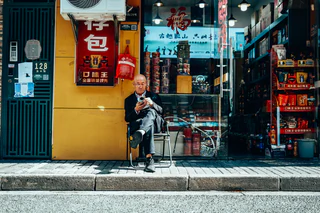 A seated man checks his phone outside a brightly coloured shop — UNSTAGED · SERIE /001 · MOMENT /004 · ARCHIVE ID S001-M004 · SOURCE L013-M003.