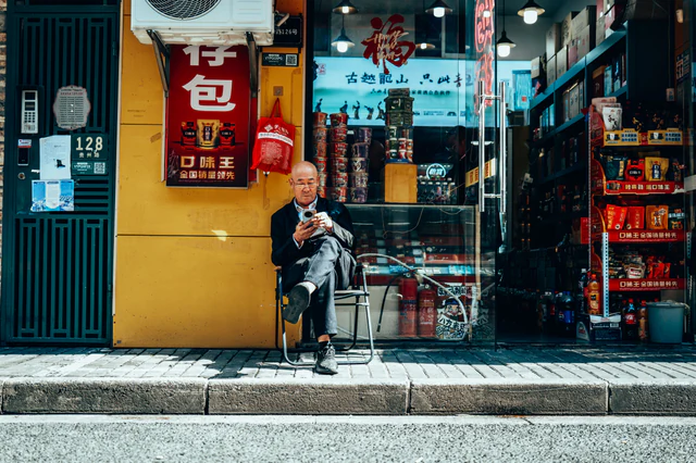 A seated man checks his phone outside a brightly coloured shop — UNSTAGED · SERIE /001 · MOMENT /004 · ARCHIVE ID S001-M004 · SOURCE L013-M003.