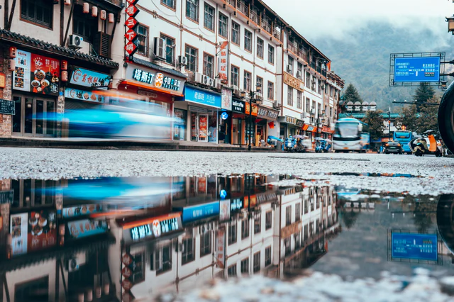 Motion-blurred vehicles pass shops reflected in a rain puddle, with misty mountains beyond — UNSTAGED · SERIE /004 · MOMENT /002 · ARCHIVE ID S004-M002 · SOURCE L013-M005.