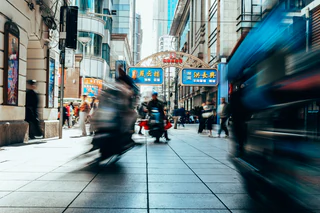 Motion-blurred scooters and pedestrians pass a sunlit row of shops with Chinese signs — UNSTAGED · LOG /013 · MOMENT /001 · ARCHIVE ID L013-M001.